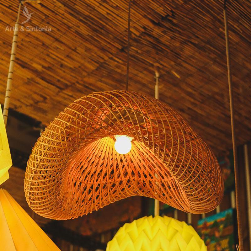 Lustre de rattan em formato fluído posicionado em sala com teto de madeira e decoração rústica.