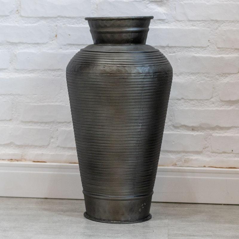 Vaso tipo urna em metal onix escuro com textura de linhas horizontais em relevo sobre fundo de parede branca.