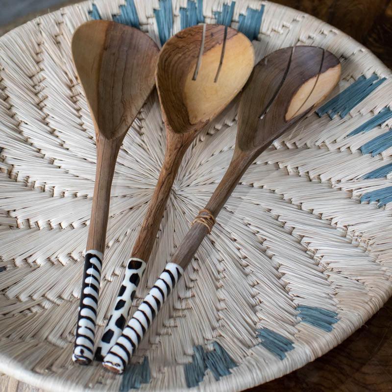 Utensílio de cozinha africano de 33cm posicionado sobre um cesto trançado de fibra natural.