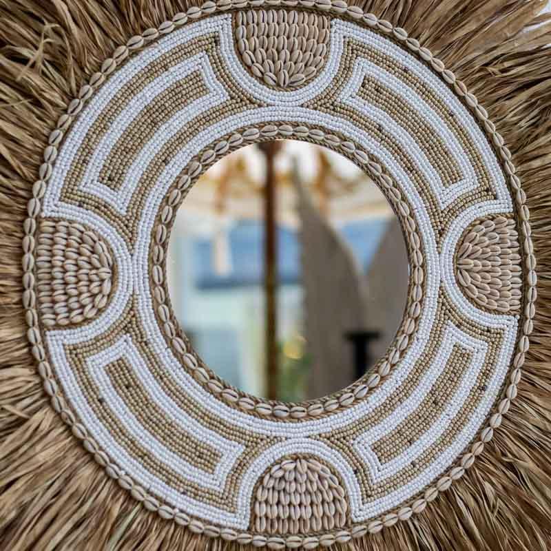 Espelho étnico redondo com moldura de franjas em fibra natural e mosaico de búzios e miçangas pretas.