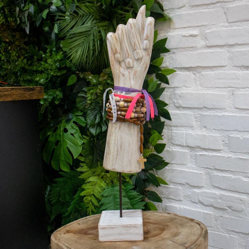 Escultura de figa artesanal em estilo luxo rústico posicionada em ambiente decorativo com plantas ao fundo.