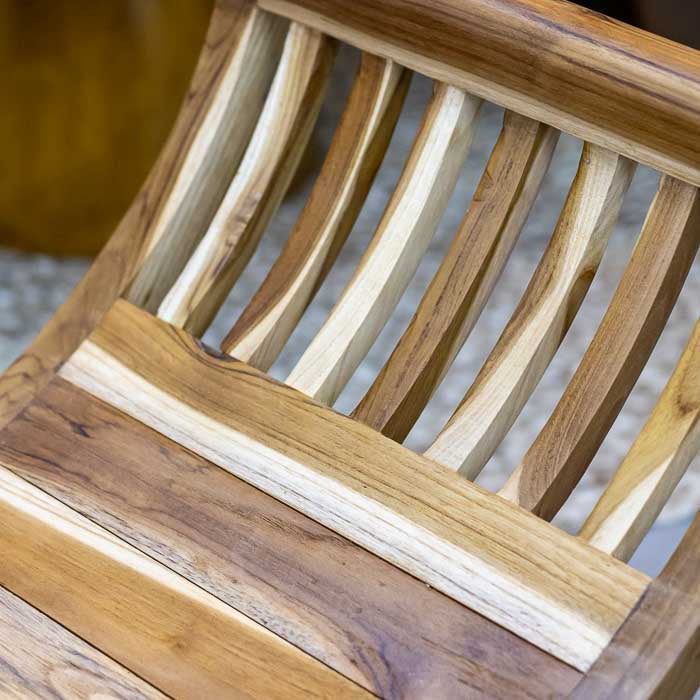 Banco de madeira teca feito manualmente em Bali com laterais curvas e base cruzada, obra que combina conforto, estética e tradição artesanal.