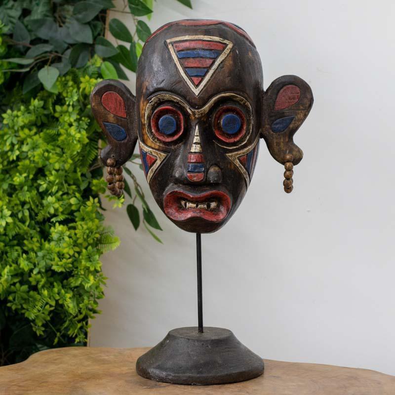 Detalhe frontal de máscara primitiva indonésia com olhos grandes circulares e brincos de contas, exposta em elegante suporte de ferro.