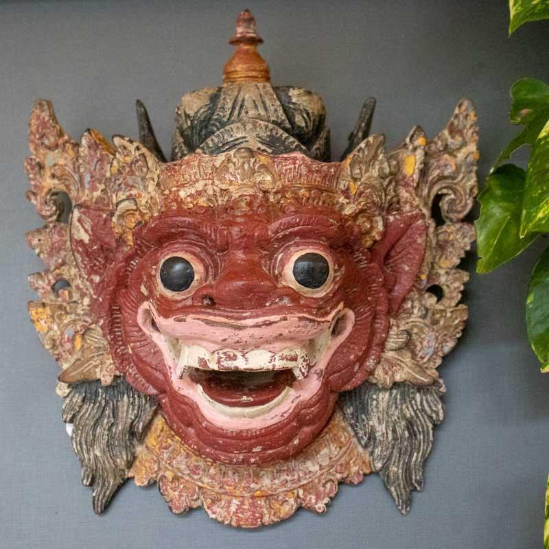 Máscara frontal de Barong em madeira entalhada com expressão feroz, olhos grandes e dentes à mostra, em estilo antik de Bali.