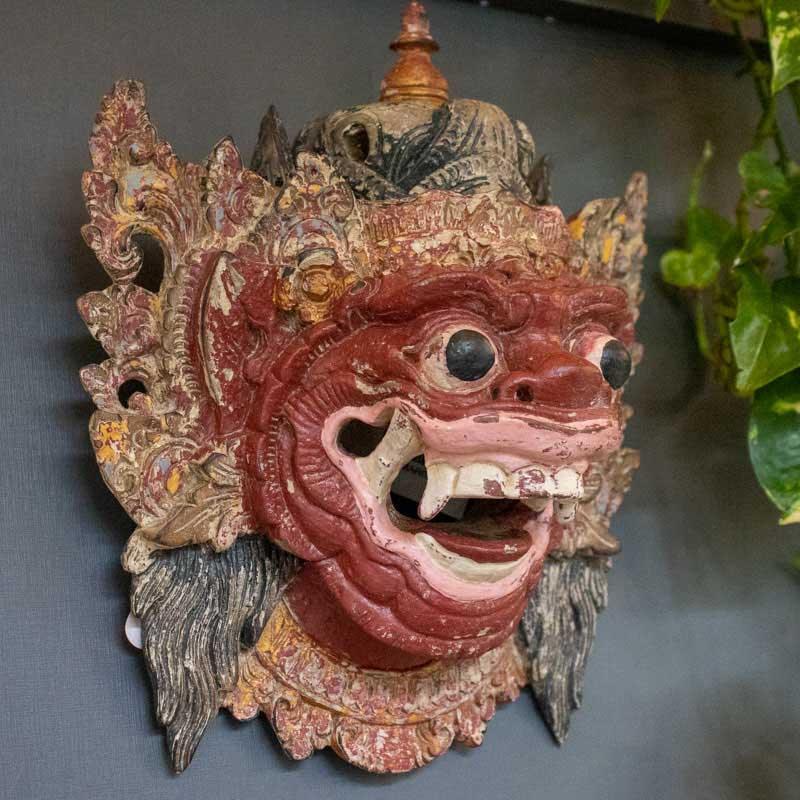 Máscara espiritual balinesa em madeira de 25cm instalada em parede cinza, destacando-se como objeto de arte étnica.