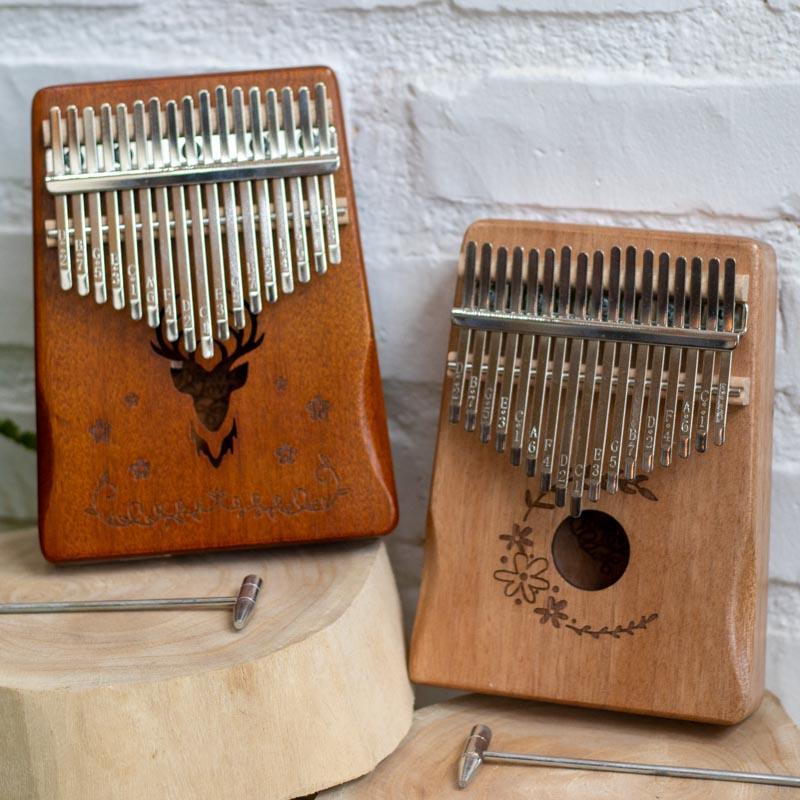 Kalimba de 17cm com martelo de afinação metálico posicionado sobre base de madeira rústica.