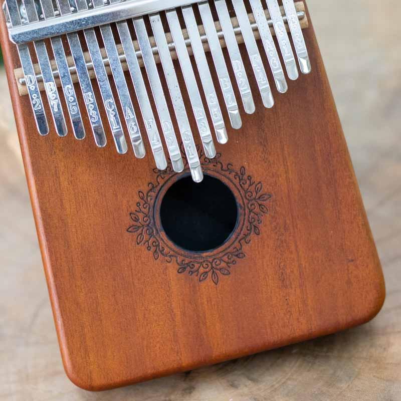 Vista frontal de Kalimba em madeira polida, destacando o design ergonômico e o entalhe artístico central.