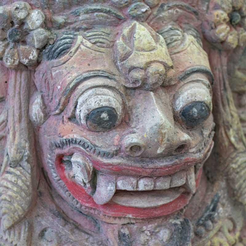 Detalhe da face da divindade Barong esculpida manualmente em pedra areia, destacando a coroa ornamental e os traços rústicos.
