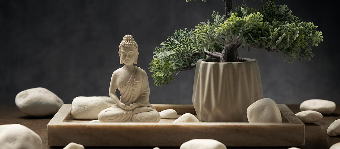 estatueta branca de Buda meditando ao lado de um bonsai com algumas pedras brancas como elementos de fundo.