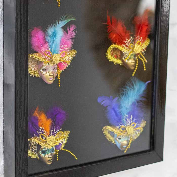 Detalhe de máscaras venezianas em tons de roxo e rosa com acabamento em glitter dourado dentro de moldura preta.