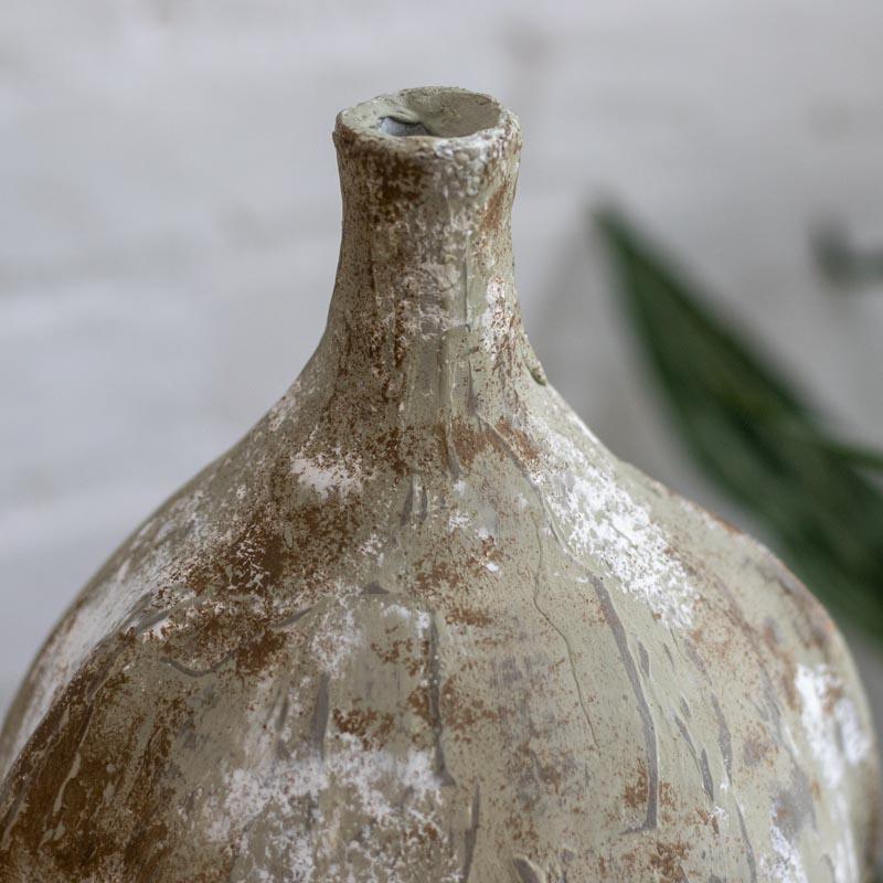 Detalhe de vaso decorativo brasileiro com pátina antik e gargalo estreito posicionado sobre móvel de madeira. 