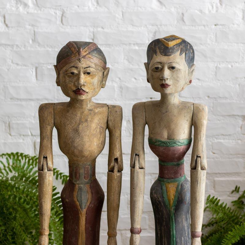 Par de esculturas Loro Blonyo em pé, feitas de madeira com braços articulados e trajes típicos javaneses em pintura patinada rústica.