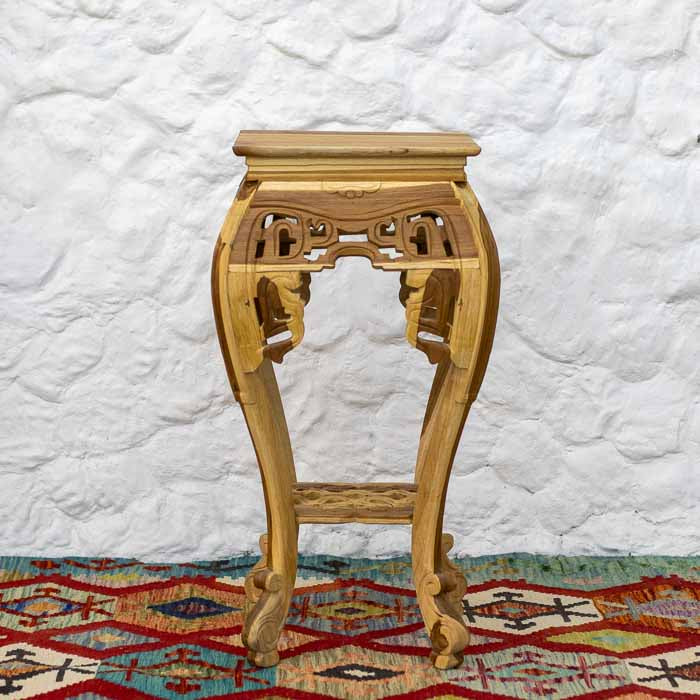 Mesa de apoio rústica em madeira natural de Bali com prateleira inferior sobre fundo de parede branca de pedra.