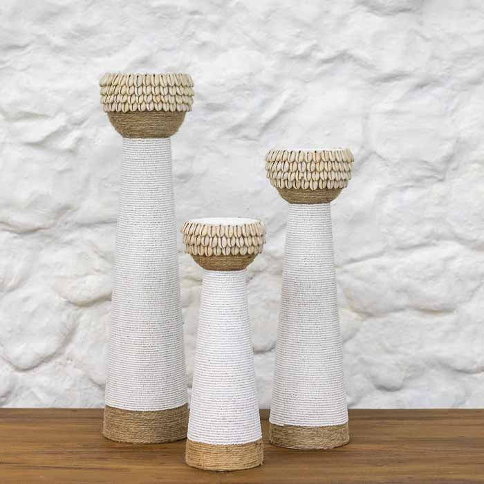 Trio de castiçais artesanais de coluna revestidos em miçangas brancas, corda natural e búzios de Bali sobre mesa.