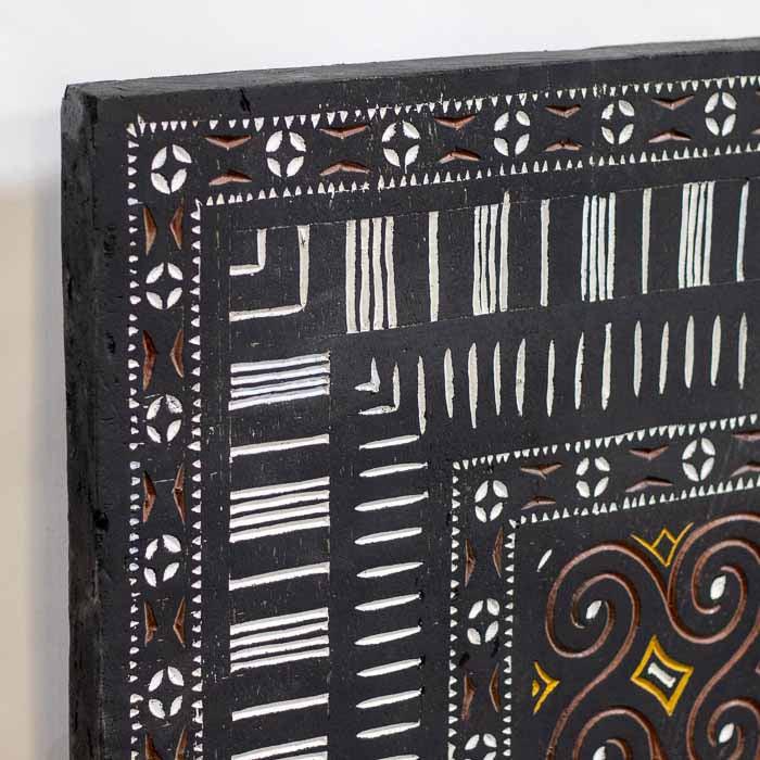 Obra de arte tribal indonésia em madeira escura com pigmentos ocre e branco instalada em parede decorativa.