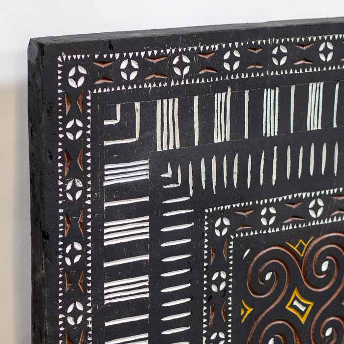 Obra de arte tribal indonésia em madeira escura com pigmentos ocre e branco instalada em parede decorativa.