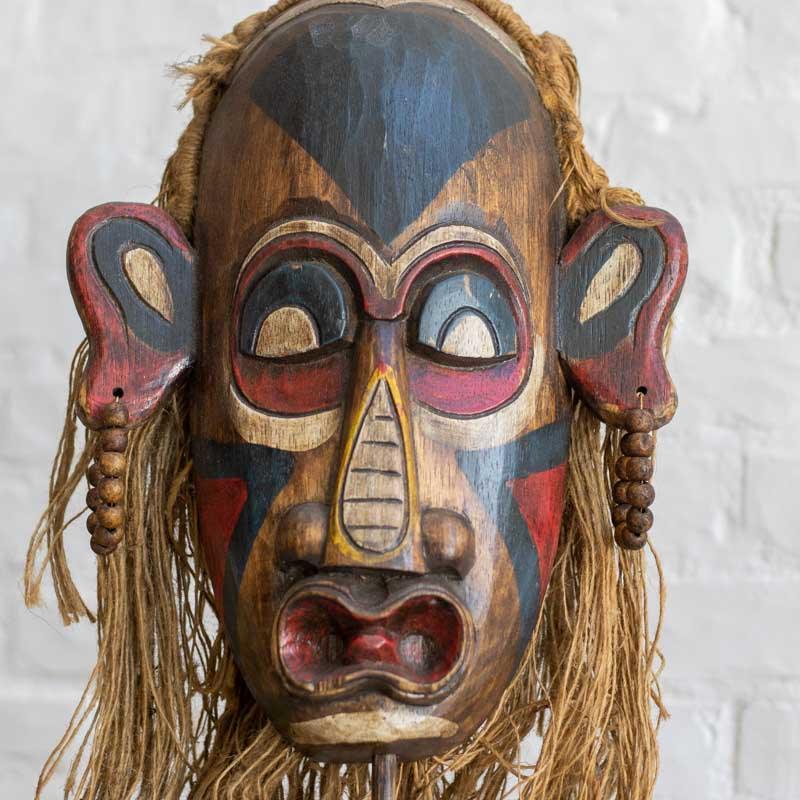 Máscara decorativa de 40cm com adornos em fibras naturais e contas de madeira, integrando arte tribal e texturas orgânicas. 