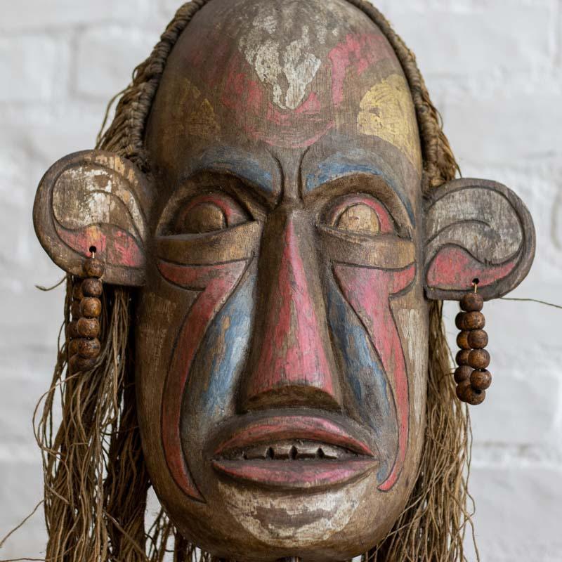 Detalhe das fibras rústicas e da pintura manual em máscara tribal de Bornéu, valorizando o artesanato autêntico. 