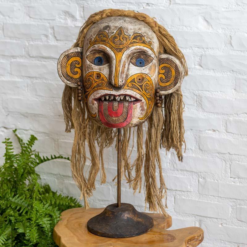 Máscara tribal de Kalimantan (Bornéu) em madeira pintada à mão, montada sobre base decorativa de madeira com haste metálica.