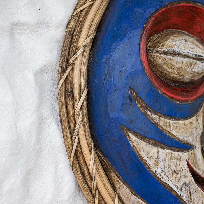 Máscara artesanal tribal feita à mão em madeira com moldura trançada em fibra natural, peça cultural de Bornéu com tons vibrantes e simbologia espiritual.