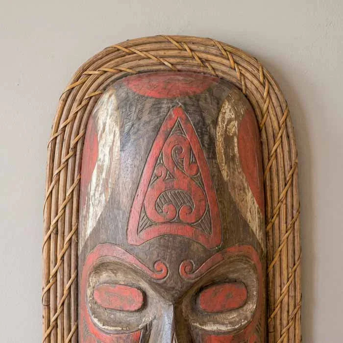 Máscara tribal Ketut de Bornéu esculpida em madeira rústica com acabamento em pintura terracota e moldura trançada de rattan.
