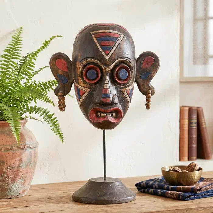 Detalhe frontal de máscara primitiva indonésia com olhos grandes circulares e brincos de contas, exposta em elegante suporte de ferro. 