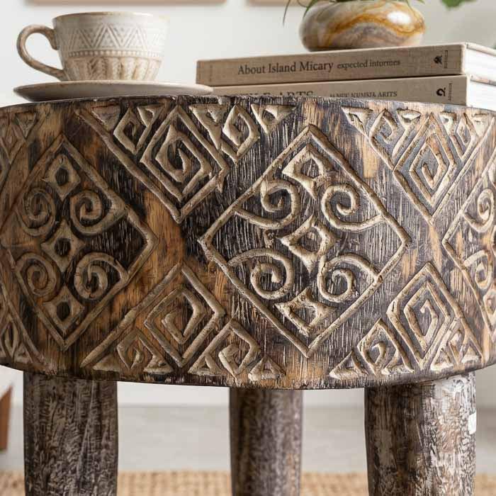 Mesa de canto artesanal em madeira maciça de Timor com entalhes tribais geométricos e acabamento em pátina branca sobre fundo escuro.