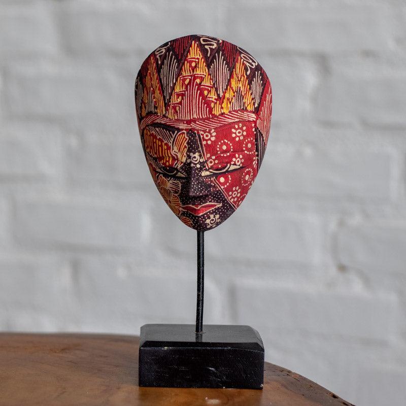 Máscara artesanal de Bali revestida com tecido Batik em tons de vermelho, preto e ocre sobre base preta. 
