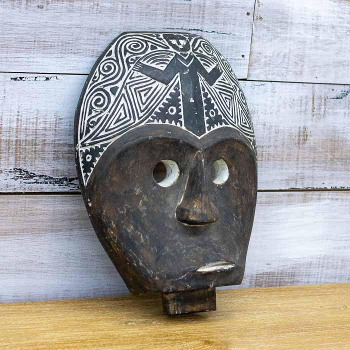 Artefacto de parede étnico em madeira escura maciça posicionado em ambiente de decoração rústica com elementos naturais e iluminação suave.