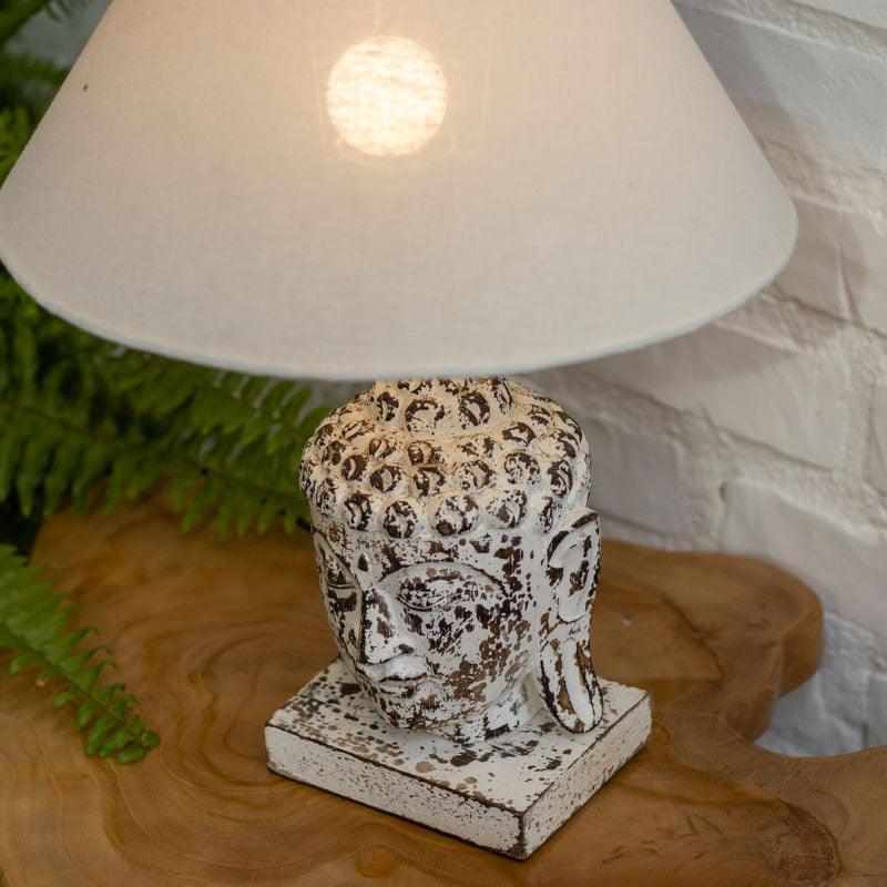 Detalhe da face serena de Buda na base de madeira de uma luminária de mesa, realçada por texturas rústicas e pátina antik de Bali.