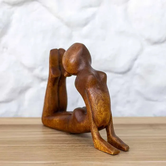 Escultura artesanal da yogini na postura Raja Bhujangasana em madeira maciça sobre mesa de tom escuro.