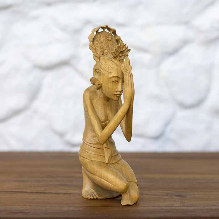 Escultura artesanal de mulher balinesa ajoelhada em oração com mãos unidas em Anjali Mudra sobre mesa de madeira.