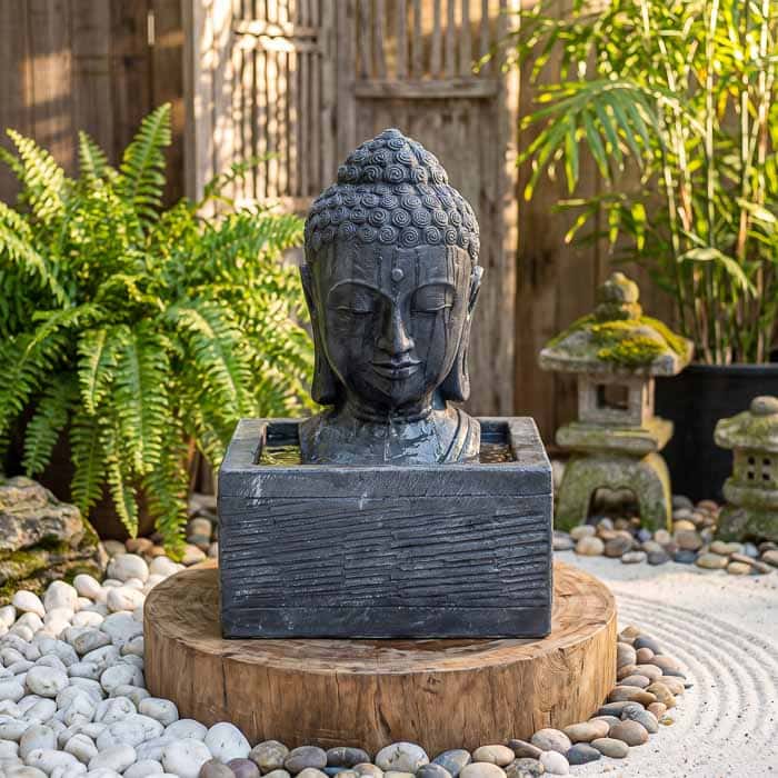 Fonte de água em formato de cabeça de Buda esculpida em cimento preto com fluxo de água superior.