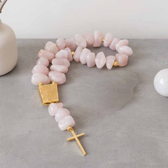 Colar de mesa terço com pedras naturais de quartzo rosa polido e cruz metálica dourada sobre bancada cinza.